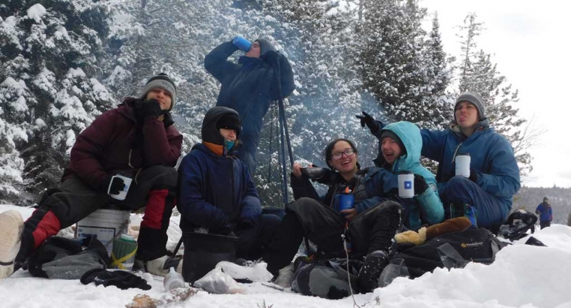 A group of people wearing heavy snow gear smile at the camera while drinking or holding mugs in front of a campfire in the snow.